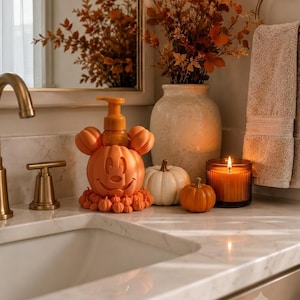 May include: An orange soap dispenser shaped like a pumpkin with mouse ears, featuring a carved face and a base decorated with small pumpkins. The dispenser is placed on a marble countertop alongside a white pumpkin, a small orange pumpkin, and a candle.