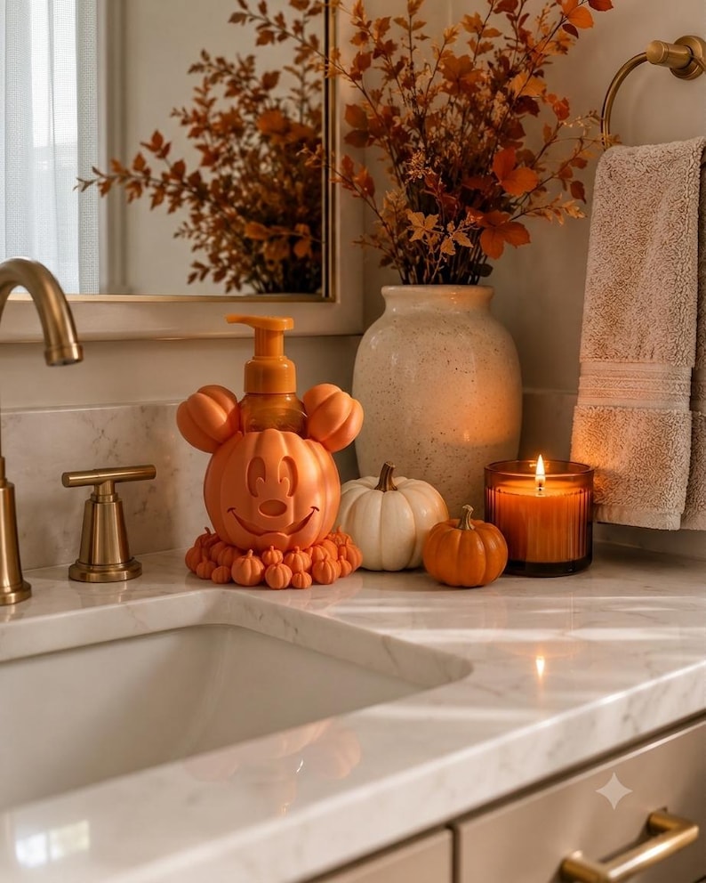 May include: An orange soap dispenser shaped like a pumpkin with mouse ears, featuring a carved face and a base decorated with small pumpkins. The dispenser is placed on a marble countertop alongside a white pumpkin, a small orange pumpkin, and a candle.