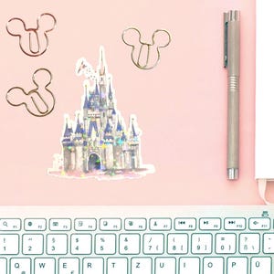 May include: A pink desk setup featuring a sticker of a fairytale castle with blue and purple accents, along with three Mickey Mouse-shaped paper clips. A silver pen and a white notepad are also present, with a keyboard in the foreground.