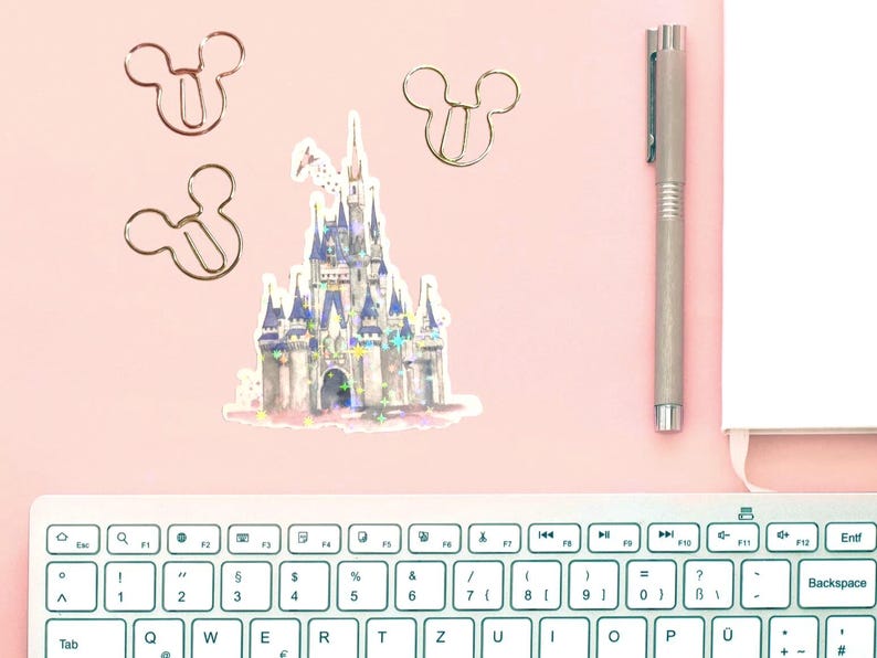 May include: A pink desk setup featuring a sticker of a fairytale castle with blue and purple accents, along with three Mickey Mouse-shaped paper clips. A silver pen and a white notepad are also present, with a keyboard in the foreground.