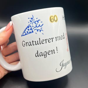 Peut inclure: Tasse en céramique blanche avec l'inscription "Gratulerer med dagen!" en noir, un motif de cotillon bleu et blanc et le chiffre 60 en or. Un motif de verre à champagne rouge et le mot "Joyeux" sont également visibles.