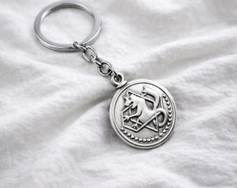 May include: Silver-colored keychain featuring a circular pendant with a detailed relief design. The pendant depicts a stylized creature within a geometric frame, surrounded by a laurel wreath. The keychain includes a ring and chain.