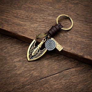 Puede incluir: Un llavero de estilo vintage con un colgante de punta de flecha de color dorado con detalles intrincados. El llavero incluye una correa de cuero marrón, un anillo de metal y dos pequeños dijes. El fondo es una superficie de madera.