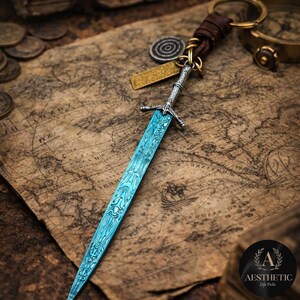 May include: A miniature sword keychain with a blue blade and silver hilt. The keychain features a leather strap and metal ring. The sword is detailed with a patterned design. The background includes a vintage map and coins.