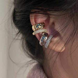May include: A set of four ear cuffs worn on the cartilage. Two cuffs feature a gold-toned hammered metal texture, while the other two are silver-toned bands encrusted with small, clear, sparkling stones arranged in a dense, pavé-style pattern.