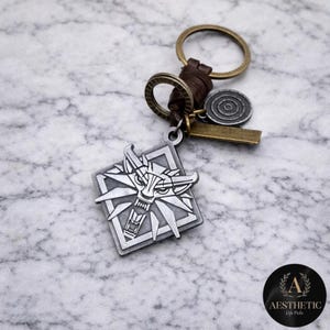 May include: A silver-colored keychain featuring a wolf head emblem within a square frame. The keychain includes a leather strap, bronze-colored rings, and a small circular charm. The background is a marble-patterned surface.