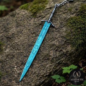May include: A detailed, blue sword pendant with intricate patterns on the blade. The sword has a silver-colored hilt and is attached to a chain. The pendant is displayed on a textured rock surface, with a background of green moss and foliage.
