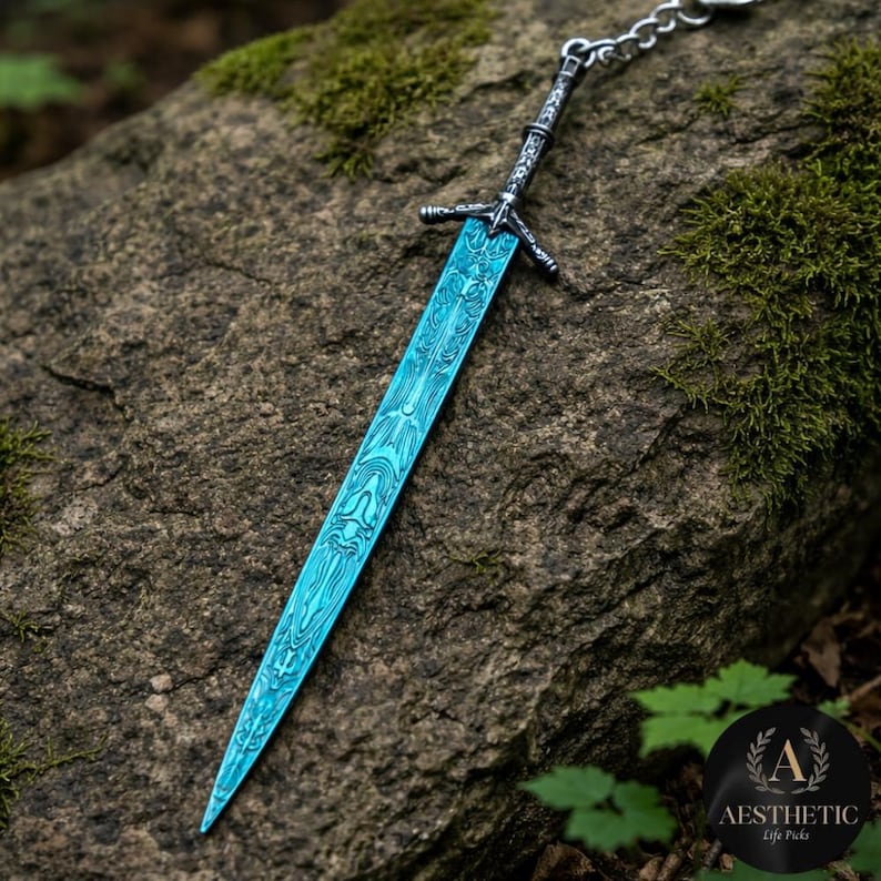 May include: A detailed, blue sword pendant with intricate patterns on the blade. The sword has a silver-colored hilt and is attached to a chain. The pendant is displayed on a textured rock surface, with a background of green moss and foliage.