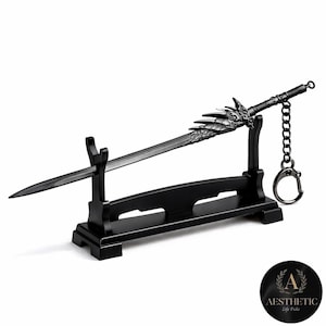 Puede incluir: Una espada negra en miniatura con intrincados detalles de dragón, exhibida en un soporte negro a juego. La espada incluye un llavero. El diseño general es elegante y gótico, con un enfoque en la empuñadura ornamentada de la hoja.