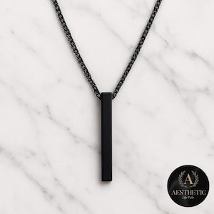 Puede incluir: Un collar con un colgante de barra vertical negro. El colgante rectangular cuelga de una cadena negra a juego. El collar se muestra sobre una superficie de mármol blanco y gris. El logotipo "Aesthetic Life Picks" está en la esquina inferior derecha.