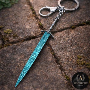 May include: A detailed keychain featuring a miniature sword with a teal blade and ornate silver handle. The blade has a unique patterned design. The keychain includes a clasp and a ring for attaching to keys or bags.