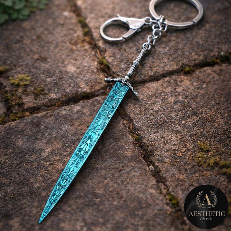 May include: A detailed keychain featuring a miniature sword with a teal blade and ornate silver handle. The blade has a unique patterned design. The keychain includes a clasp and a ring for attaching to keys or bags.