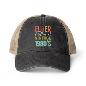 May include: A black baseball cap with a tan mesh back and a distressed finish. The front features the text ELDER MILLENNIAL VINTAGE 1980'S in a multi-colored, textured font with red, orange, yellow, and light blue lettering on the black fabric.