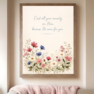 May include: A framed print with the text "Cast all your anxiety on Him, because He cares for you. 1 Peter 5:7." The artwork has a floral border with pink, blue, and white flowers. The frame is a light brown colour.