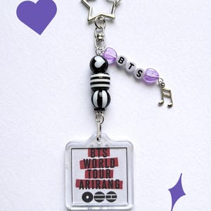 May include: A keychain featuring a star-shaped clasp, black and white striped and solid beads, and a clear tag with the text "BTS WORLD TOUR ARIRANG". The keychain also includes purple heart-shaped beads and a musical note charm.