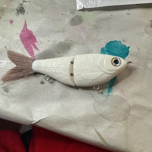 May include: A white fishing lure shaped like a fish, with a segmented body and a brown tail. The lure has a detailed scale pattern and a reflective eye. It is designed to attract fish.