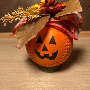 Puede incluir: Decoración de calabaza naranja hecha a mano con un diseño cosido y una cara sonriente de jack-o'-lantern. Está adornada con una cinta a cuadros, trigo seco y flores de otoño. La calabaza se asienta sobre una base verde.