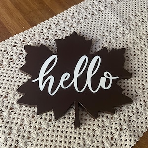 Peut inclure: Décoration en forme de feuille d'érable en bois marron foncé avec le mot "hello" en écriture blanche. La feuille est posée sur un chemin de table en macramé crème, avec une table en bois en arrière-plan.