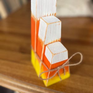 Può includere: Decorazione in legno a forma di caramella di mais. I blocchi impilati sono dipinti in giallo, arancione e bianco e legati con spago. I bordi dei blocchi sono leggermente usurati. Decorazione a tema Halloween.