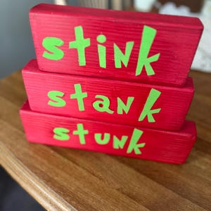 Könnte beinhalten: Drei rote Holzblöcke, die übereinander gestapelt sind. Jeder Block trägt ein anderes Wort in leuchtend grünen Buchstaben: "stink", "stank" und "stunk". Die Blöcke liegen auf einer Holzoberfläche. Ein humorvolles Dekorationsstück.