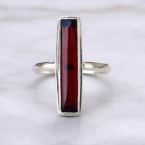 Peut inclure: Une bague en argent avec une pierre précieuse rectangulaire rouge foncé. La pierre est sertie dans une lunette en argent, et la bande est en argent poli simple. La bague est présentée sur une surface en marbre blanc.