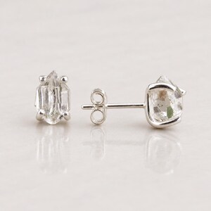 Peut inclure: Une paire de boucles d'oreilles à clous avec des cristaux bruts, transparents et hexagonaux, sertis dans des griffes en métal argenté. Une boucle est vue de face, l'autre de profil, montrant la tige et le fermoir papillon en métal.