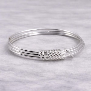 May include: A silver-colored bangle bracelet. The bracelet is made of multiple thin, parallel wires, with a coiled design at the center. The bracelet has a shiny, reflective surface and is set against a light gray background.
