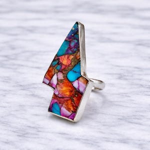 Op de afbeelding: Een zilveren ring met een grote, kleurrijke steen. De steen is een mix van turquoise, paars, oranje en goud. De ring heeft een unieke, geometrische vorm.