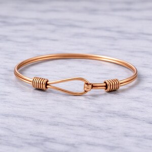 Puede incluir: Una pulsera de brazalete de oro rosa. La pulsera tiene un cierre de gancho y ojo. Los extremos de la pulsera están envueltos con alambre. La pulsera está sobre una superficie gris claro.