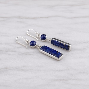 May include: A pair of drop earrings featuring a silver hook, a small round lapis lazuli stone, and a rectangular lapis lazuli pendant. The deep blue stones contain natural gold-colored flecks, set within polished silver frames for a clean look.