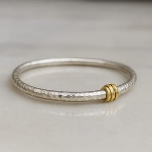 May include: A silver bangle bracelet with a hammered texture. The bracelet has a gold-coloured accent with three stacked rings. The jewellery is displayed on a white surface.