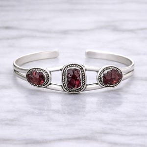 May include: A silver cuff bracelet featuring three raw, deep red garnet stones. The stones are set in oval and rectangular frames with a twisted rope design. The bracelet has a simple, open design.