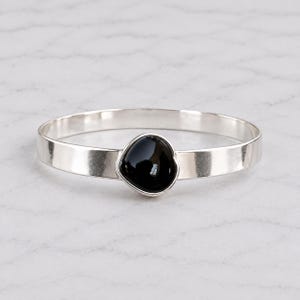 Op de afbeelding: Een gepolijste zilveren armband met een enkele, gladde, druppelvormige zwarte steen in het midden. De metalen band heeft een reflecterende, hoogglanzende afwerking en een consistente breedte, wat zorgt voor een strak en eenvoudig cirkelvormig silhouet.