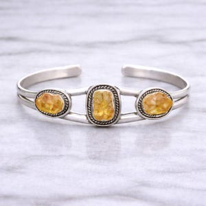 Op de afbeelding: Zilveren manchetarmband met drie ruwe citrienstenen. De stenen zijn gezet in zilveren bezels met een gedraaid touwontwerp. De armband heeft een open ontwerp. De citrienstenen zijn ovaal van vorm.