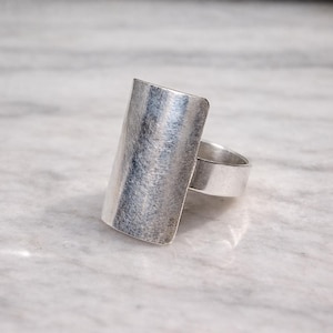 Könnte beinhalten: Ein silberfarbener Ring mit rechteckiger Oberfläche. Der Ring hat eine gebürstete Metallstruktur und ein breites Band. Die rechteckige Fläche ist ca. 3,8 cm lang und 1,9 cm breit, was ihm einen kühnen, modernen Look verleiht.