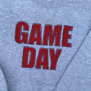 Könnte beinhalten: Ein hellgraues Sweatshirt mit dem Text "GAME DAY" in großen, roten Blockbuchstaben. Die Buchstaben haben ein gepunktetes Muster. Das Sweatshirt hat einen Rundhalsausschnitt und lange Ärmel.