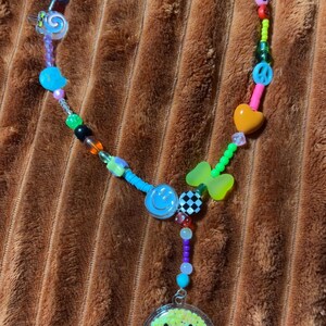 May include: A colorful beaded necklace with various charms, including a smiley face pendant, peace signs, hearts, and stars. The necklace features a mix of vibrant beads in blue, green, pink, and orange. The necklace is displayed on a brown, textured surface.