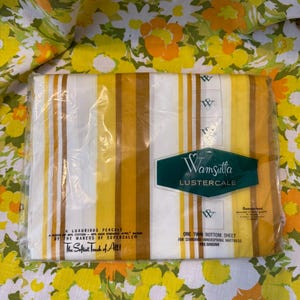 May include: A packaged twin bottom sheet with vertical stripes in shades of yellow, white, and brown. The sheet is labeled "Wamsutta Lustercale" and is pre-shrunk. The background features a floral pattern in yellow, orange, and white.