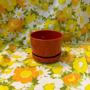 Puede incluir: Una vibrante maceta de cerámica naranja con un platillo a juego. La maceta es cilíndrica con un acabado liso y brillante. El fondo es un estampado floral en amarillo, naranja y blanco.
