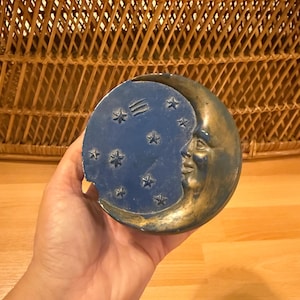 Könnte beinhalten: Eine dekorative Schale mit einem himmlischen Thema, die einen Halbmond und Sterne darstellt. Die Schale ist in Blau und Gold gehalten, mit einem lächelnden Mondgesicht und Sternen. Die Schale wird in einer Hand gehalten.