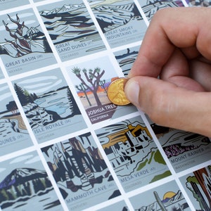 May include: A scratch-off map featuring illustrations of U.S. National Parks. The map is light gray with colorful illustrations. A hand holds a penny over the Joshua Tree National Park square. The text "JOSHUA TREE CALIFORNIA" is visible.
