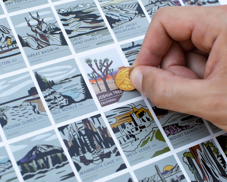 May include: A scratch-off map featuring illustrations of U.S. National Parks. The map is light gray with colorful illustrations. A hand holds a penny over the Joshua Tree National Park square. The text "JOSHUA TREE CALIFORNIA" is visible.