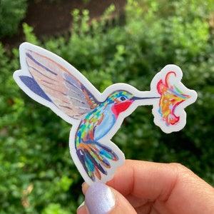 May include: A colorful sticker featuring a hummingbird with a long beak, holding a flower. The hummingbird has blue, green, red, and yellow feathers. The flower is a mix of orange, yellow, and red. The sticker is on a white background.