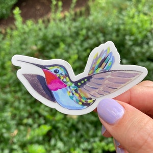 May include: A colorful hummingbird sticker with a white border. The bird features a red throat, blue body, and multi-colored wings. The sticker is held against a blurred green background.