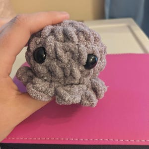 Puede incluir: Un pequeño pulpo de ganchillo gris hecho a mano con grandes ojos negros. El peluche tiene ocho tentáculos y una superficie texturizada. El pulpo se sostiene en una mano y descansa sobre una superficie rosa.