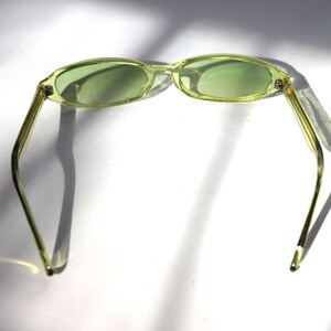 K&ouml;nnte beinhalten: Eine ovale Sonnenbrille mit hellgr&uuml;ner T&ouml;nung. Die Fassungen und B&uuml;gel sind hellgelb-gr&uuml;n. Die Sonnenbrille liegt vor einem wei&szlig;en Hintergrund mit Schatten.