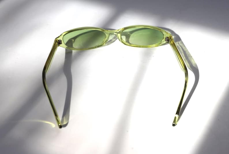 K&ouml;nnte beinhalten: Eine ovale Sonnenbrille mit hellgr&uuml;ner T&ouml;nung. Die Fassungen und B&uuml;gel sind hellgelb-gr&uuml;n. Die Sonnenbrille liegt vor einem wei&szlig;en Hintergrund mit Schatten.