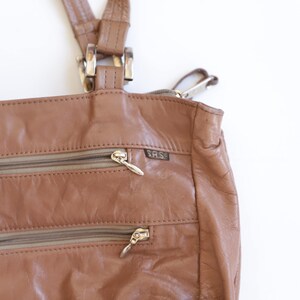 May include: A tan leather shoulder bag featuring two horizontal zippered exterior pockets with silver-toned pulls. The bag includes a small rectangular tag with the text S.A.S. attached near the top zipper. It has two leather straps with silver hardware.