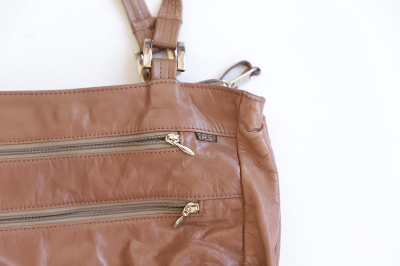 May include: A tan leather shoulder bag featuring two horizontal zippered exterior pockets with silver-toned pulls. The bag includes a small rectangular tag with the text S.A.S. attached near the top zipper. It has two leather straps with silver hardware.