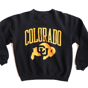May include: Black crew neck sweatshirt with the word "COLORADO" in gold block letters across the chest. Below the word "COLORADO" is a gold graphic of a buffalo. The sweatshirt is made of a soft material.